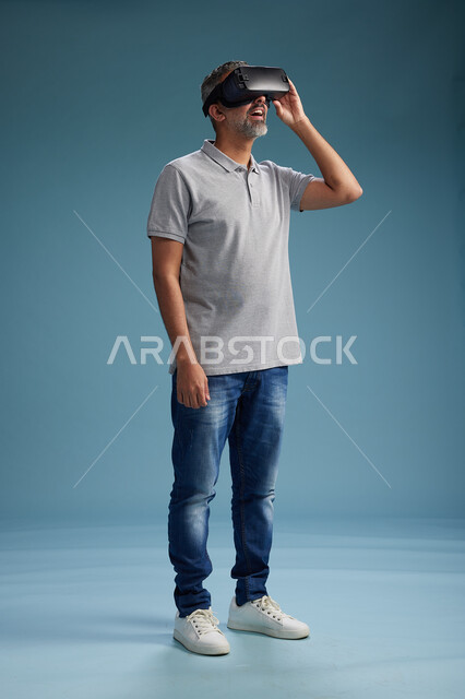 رجل عربي سعودي خليجي يرتدي الزي الكاجوال يستخدم نظارة الواقع الافتراضي, مشاهدة الافلام والفيديوز المسليه عبر الفي ار, كامل الجسم, خلفيه زرقاء