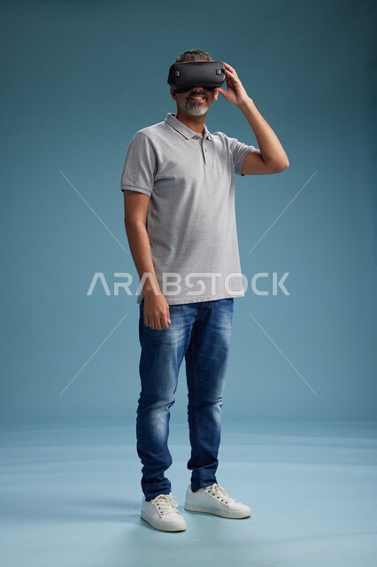 رجل عربي سعودي خليجي يرتدي الزي الكاجوال يستخدم نظارة الواقع الافتراضي, مشاهدة الافلام والفيديوز المسليه عبر الفي ار, كامل الجسم, خلفيه زرقاء