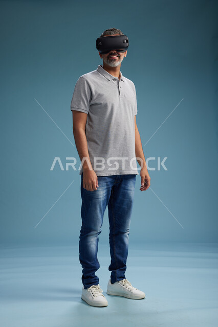 A Saudi Arabian Gulf Arab man wearing casual attire is using virtual reality glasses, watching movies and entertaining videos through VR, full body, with a blue background.