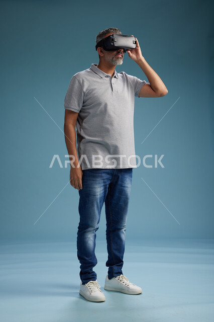 رجل عربي سعودي خليجي يرتدي الزي الكاجوال يستخدم نظارة الواقع الافتراضي, مشاهدة الافلام والفيديوز المسليه عبر الفي ار, كامل الجسم, خلفيه زرقاء