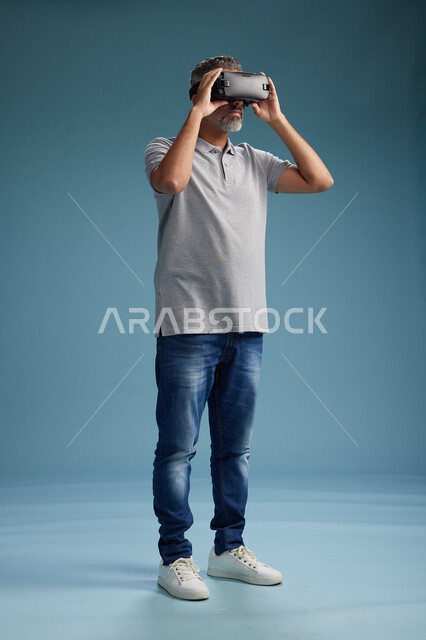 رجل عربي سعودي خليجي يرتدي الزي الكاجوال يستخدم نظارة الواقع الافتراضي, مشاهدة الافلام والفيديوز المسليه عبر الفي ار, كامل الجسم, خلفيه زرقاء