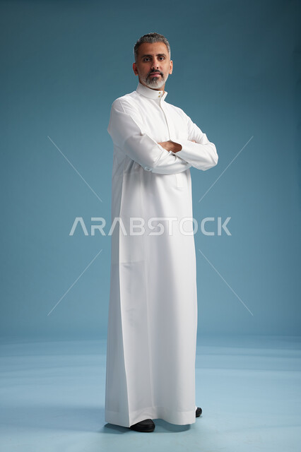 رجل عربي  سعودي خليجي يرتدي ثوب ابيض  بأيدي متقاطعه, النظر الى الكاميرا, الوقوف بثقه, كامل الجسم, خلفيه زرقاء
