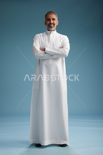 A Saudi Arabian Gulf Arab man wearing a white thobe, with his hands crossed, looking at the camera, standing confidently, full body, blue background.