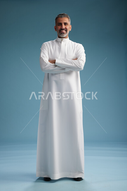 A Saudi Arabian Gulf Arab man wearing a white thobe, with his hands crossed, looking at the camera, standing confidently, full body, blue background.