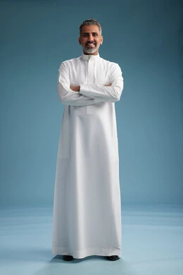رجل عربي  سعودي خليجي يرتدي ثوب ابيض  بأيدي متقاطعه, النظر الى الكاميرا, الوقوف بثقه, كامل الجسم, خلفيه زرقاء