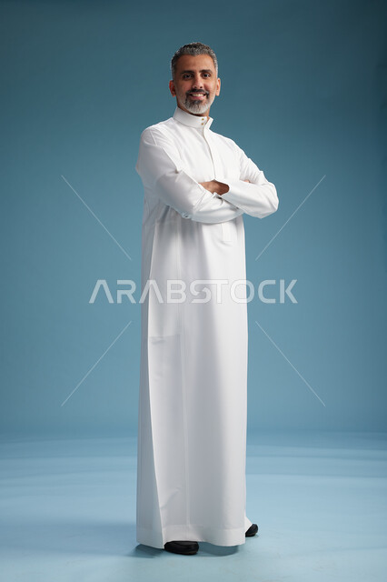 A Saudi Arabian Gulf Arab man wearing a white thobe, with his hands crossed, looking at the camera, standing confidently, full body, blue background.