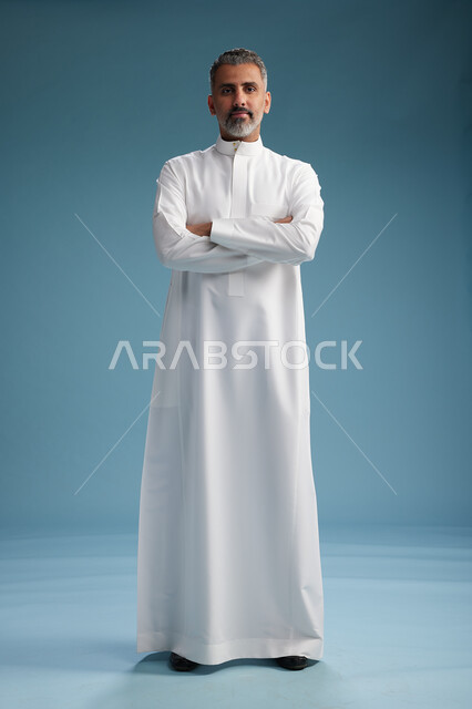 A Saudi Arabian Gulf Arab man wearing a white thobe, with his hands crossed, looking at the camera, standing confidently, full body, blue background.