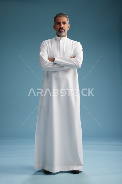 A Saudi Arabian Gulf Arab man wearing a white thobe, with his hands crossed, looking at the camera, standing confidently, full body, blue background.