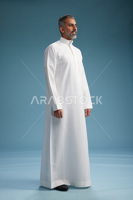 رجل عربي  سعودي خليجي يرتدي ثوب ابيض  , النظر الى شيء ما, الوقوف بثقه, كامل الجسم, خلفيه زرقاء
