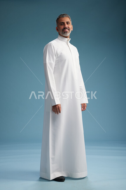 رجل عربي  سعودي خليجي يرتدي ثوب ابيض  , النظر الى شيء ما, الوقوف بثقه, كامل الجسم, خلفيه زرقاء