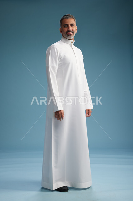 رجل عربي  سعودي خليجي يرتدي ثوب ابيض  , النظر الى شيء ما, الوقوف بثقه, كامل الجسم, خلفيه زرقاء