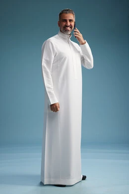 A Saudi Arabian man from the Gulf region, wearing a white thobe, makes a phone call with various gestures and movements, communicating with friends via mobile phone, full body, blue background.