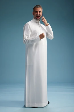 A Saudi Arabian man from the Gulf region, wearing a white thobe, makes a phone call with various gestures and movements, communicating with friends via mobile phone, full body, blue background.