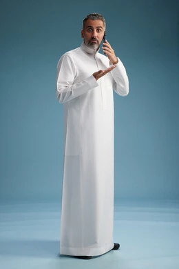 A Saudi Arabian man from the Gulf region, wearing a white thobe, makes a phone call with various gestures and movements, communicating with friends via mobile phone, full body, blue background.