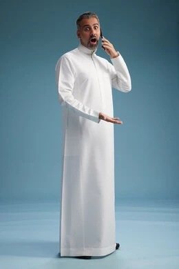 A Saudi Arabian man from the Gulf region, wearing a white thobe, makes a phone call with various gestures and movements, communicating with friends via mobile phone, full body, blue background.