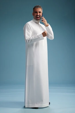 A Saudi Arabian man from the Gulf region, wearing a white thobe, makes a phone call with various gestures and movements, communicating with friends via mobile phone, full body, blue background.