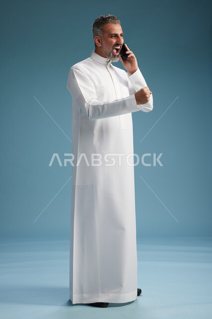 رجل  عربي سعودي خليجي يرتدي الثوب الابيض يجري مكالمة هاتفية بايماءات وحركات متنوعه, التواصل مع الاصدقاء عبر الجوال, كامل الجسم, خلفيه زرقاء