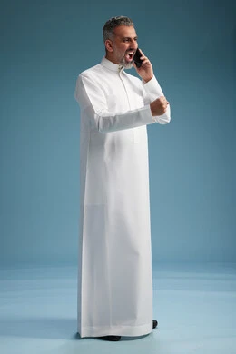 A Saudi Arabian man from the Gulf region, wearing a white thobe, makes a phone call with various gestures and movements, communicating with friends via mobile phone, full body, blue background.
