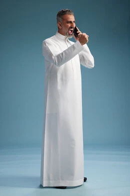 A Saudi Arabian man from the Gulf region, wearing a white thobe, makes a phone call with various gestures and movements, communicating with friends via mobile phone, full body, blue background.