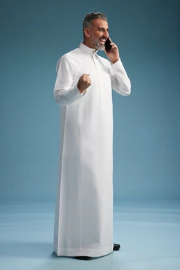 رجل  عربي سعودي خليجي يرتدي الثوب الابيض يجري مكالمة هاتفية بايماءات وحركات متنوعه, التواصل مع الاصدقاء عبر الجوال, كامل الجسم, خلفيه زرقاء