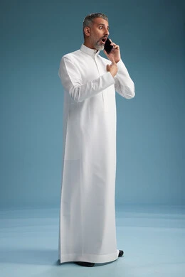 رجل  عربي سعودي خليجي يرتدي الثوب الابيض يجري مكالمة هاتفية بايماءات وحركات متنوعه, التواصل مع الاصدقاء عبر الجوال, كامل الجسم, خلفيه زرقاء