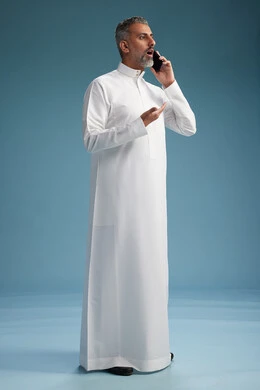 رجل  عربي سعودي خليجي يرتدي الثوب الابيض يجري مكالمة هاتفية بايماءات وحركات متنوعه, التواصل مع الاصدقاء عبر الجوال, كامل الجسم, خلفيه زرقاء