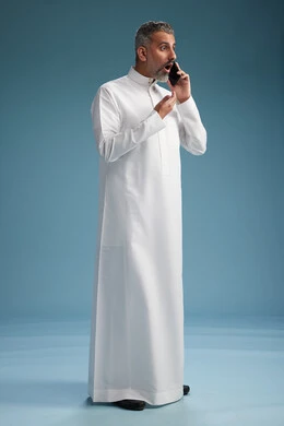 A Saudi Arabian man from the Gulf region, wearing a white thobe, makes a phone call with various gestures and movements, communicating with friends via mobile phone, full body, blue background.