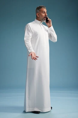 رجل  عربي سعودي خليجي يرتدي الثوب الابيض يجري مكالمة هاتفية بايماءات وحركات متنوعه, التواصل مع الاصدقاء عبر الجوال, كامل الجسم, خلفيه زرقاء