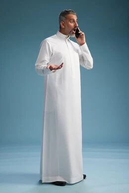 رجل  عربي سعودي خليجي يرتدي الثوب الابيض يجري مكالمة هاتفية بايماءات وحركات متنوعه, التواصل مع الاصدقاء عبر الجوال, كامل الجسم, خلفيه زرقاء