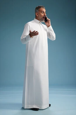 A Saudi Arabian man from the Gulf region, wearing a white thobe, makes a phone call with various gestures and movements, communicating with friends via mobile phone, full body, blue background.