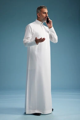 رجل  عربي سعودي خليجي يرتدي الثوب الابيض يجري مكالمة هاتفية بايماءات وحركات متنوعه, التواصل مع الاصدقاء عبر الجوال, كامل الجسم, خلفيه زرقاء