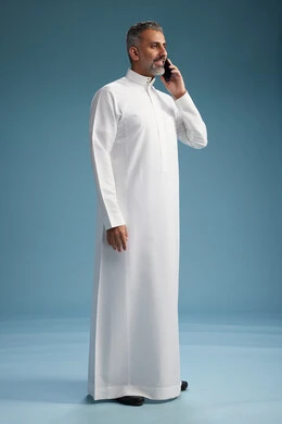 A Saudi Arabian man from the Gulf region, wearing a white thobe, makes a phone call with various gestures and movements, communicating with friends via mobile phone, full body, blue background.