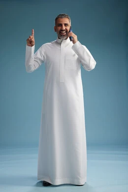 A Saudi Arabian man from the Gulf region, wearing a white thobe, makes a phone call with various gestures and movements, communicating with friends via mobile phone, full body, blue background.