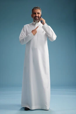 A Saudi Arabian man from the Gulf region, wearing a white thobe, makes a phone call with various gestures and movements, communicating with friends via mobile phone, full body, blue background.