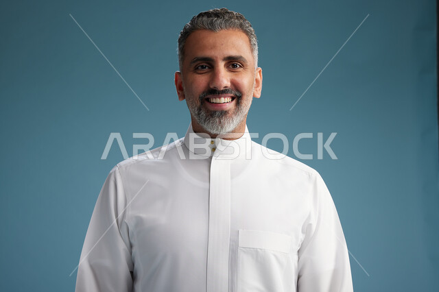 رجل عربي  سعودي خليجي يرتدي ثوب ابيض  بأيدي متقاطعه, النظر الى الكاميرا, الوقوف بثقه, كامل الجسم, خلفيه زرقاء