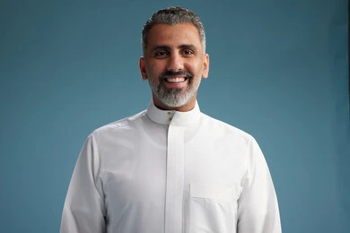 رجل عربي  سعودي خليجي يرتدي ثوب ابيض  بأيدي متقاطعه, النظر الى الكاميرا, الوقوف بثقه, كامل الجسم, خلفيه زرقاء