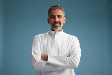 A Saudi Arabian man from the Gulf region, wearing a white thobe, with his hands crossed, looking at the camera, blue background.