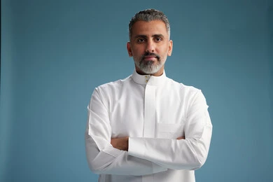A Saudi Arabian man from the Gulf region, wearing a white thobe, with his hands crossed, looking at the camera, blue background.