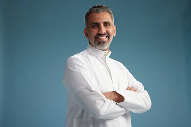 A Saudi Arabian man from the Gulf region, wearing a white thobe, with his hands crossed, looking at the camera, blue background.