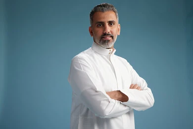 A Saudi Arabian man from the Gulf region, wearing a white thobe, with his hands crossed, looking at the camera, blue background.