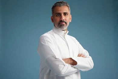 A Saudi Arabian man from the Gulf region, wearing a white thobe, with his hands crossed, looking at the camera, blue background.
