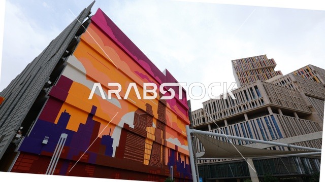 تصوير معماري لمدينة الرياض الحديثة، مبانٍ شاهقة بتصميم مستقبلي في منطقة الأعمال مركز الملك عبدالله المالي، مع عناصر فنية حضرية تضيف طابعاً إبداعياً يعكس رؤية المملكة للتطوير العمراني