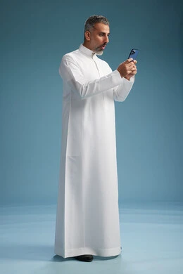 A Saudi Arabian Gulf Arab man wearing a white thobe is using a smartphone, full-body shot, blue background.