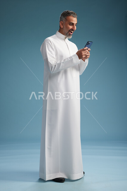 رجل  عربي سعودي خليجي يرتدي الثوب الابيض يستخدم هاتفاً ذكياً, الجسم بالطول الكامل, خلفيه زرقاء