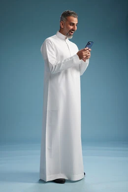 A Saudi Arabian Gulf Arab man wearing a white thobe is using a smartphone, full-body shot, blue background.