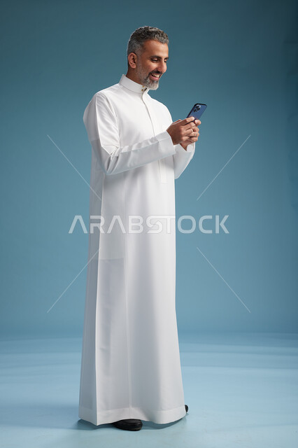 رجل  عربي سعودي خليجي يرتدي الثوب الابيض يستخدم هاتفاً ذكياً, الجسم بالطول الكامل, خلفيه زرقاء