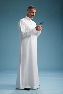 A Saudi Arabian Gulf Arab man wearing a white thobe is using a smartphone, full-body shot, blue background.
