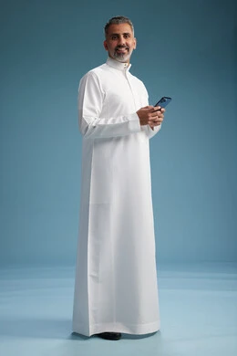 A Saudi Arabian Gulf Arab man wearing a white thobe is using a smartphone, full-body shot, blue background.