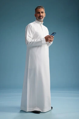 رجل  عربي سعودي خليجي يرتدي الثوب الابيض يستخدم هاتفاً ذكياً, الجسم بالطول الكامل, خلفيه زرقاء