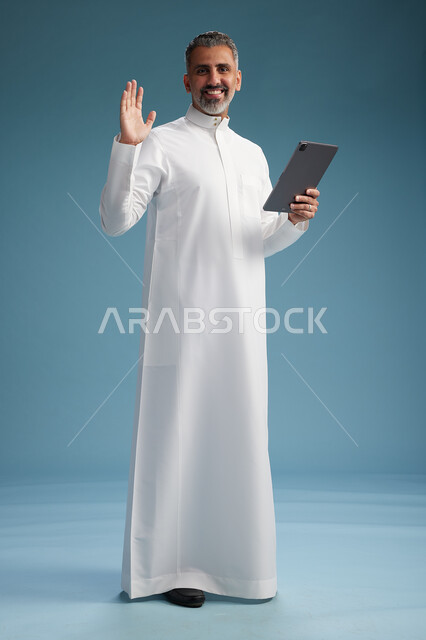 A portrait of a Saudi Arabian Gulf man wearing a white thobe using a digital tablet, full body, with a blue background.